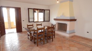 A dining area with a tiled floor, wooden table, chairs, a fireplace, and a large window with a view. - AffittaSardegna - Casa Pompia 6 (Siniscola)