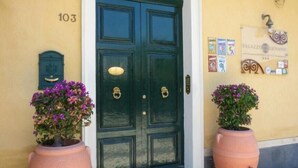 Una puerta doble verde oscuro con un picaporte de latón, flanqueada por dos macetas de terracota con flores moradas, el número 103 en la pared y un pequeño letrero con el nombre "Palazzo Giovannini".