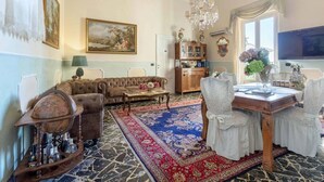 Una habitación tradicional con sofá, una mesa de comedor con sillas, una araña, un cuadro grande y un armario.