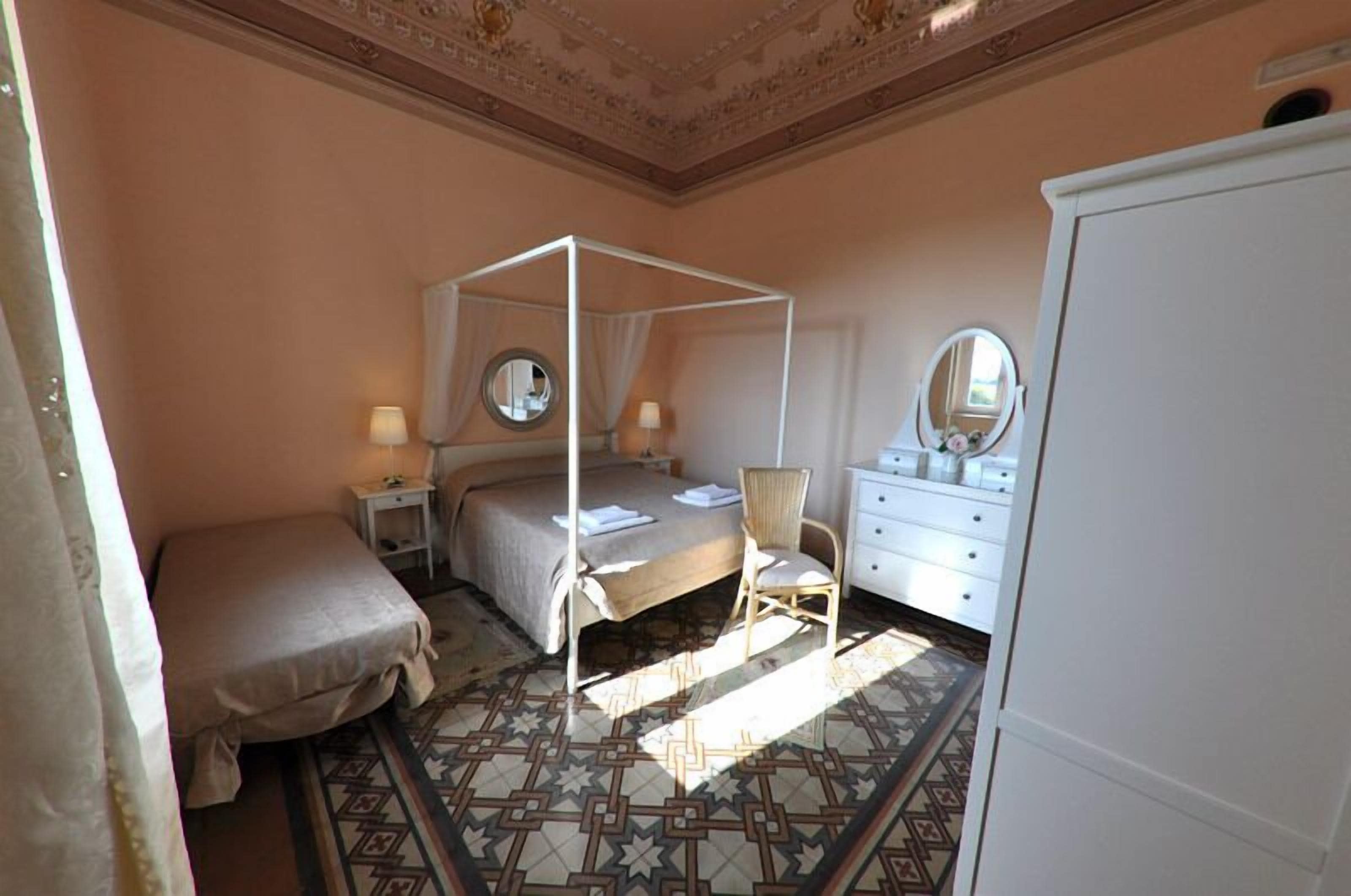 A bedroom with a four-poster bed, a bedside table with a lamp, a chair, a dresser with a mirror, and a wardrobe.
