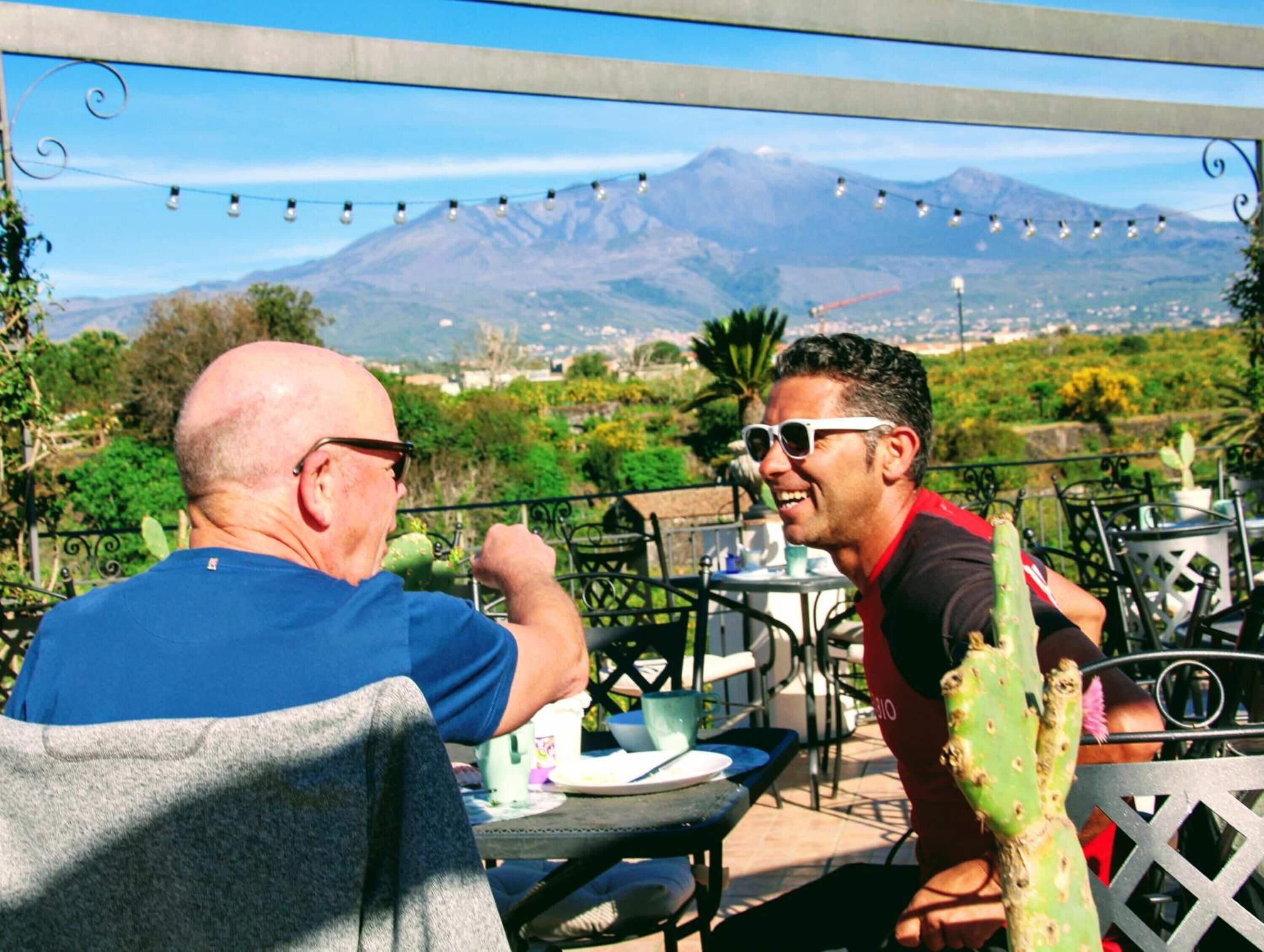 Two men seated at an outdoor table with a mountain view in the background.