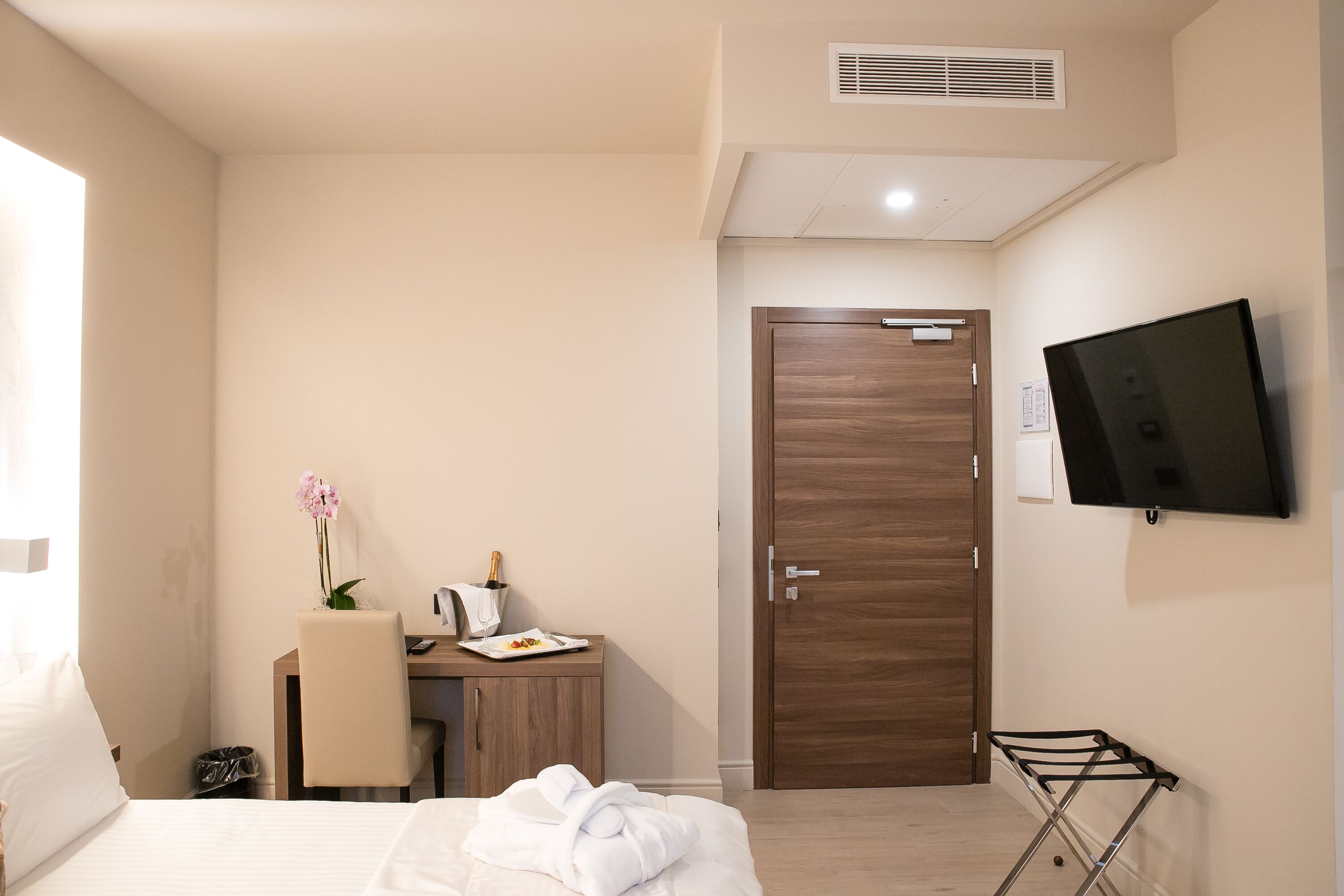 A hotel room with a bed, a desk with a chair, a TV mounted on the wall, and a wooden door.