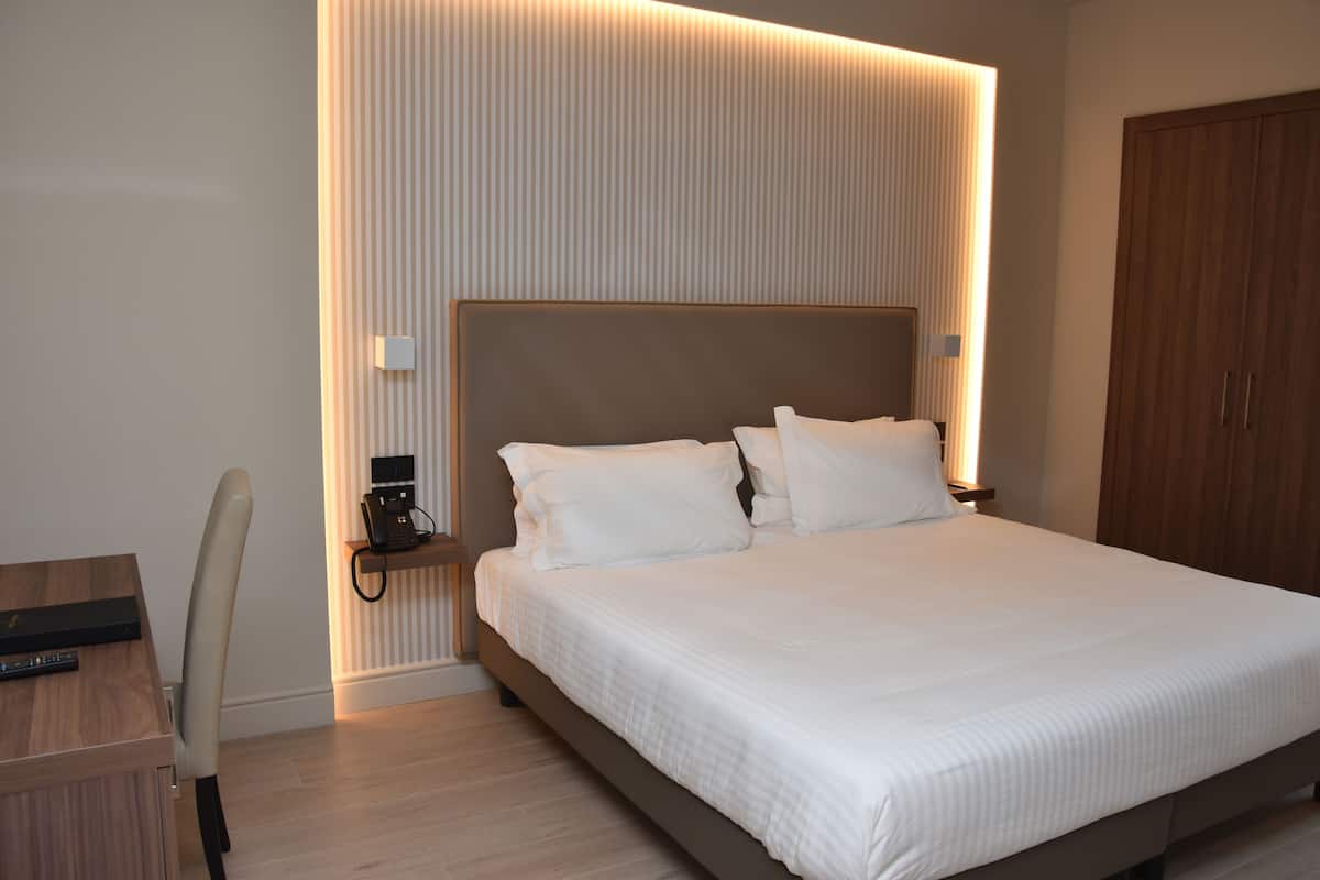 A modern hotel room with a large bed, a desk with a chair, and a wooden wardrobe.
