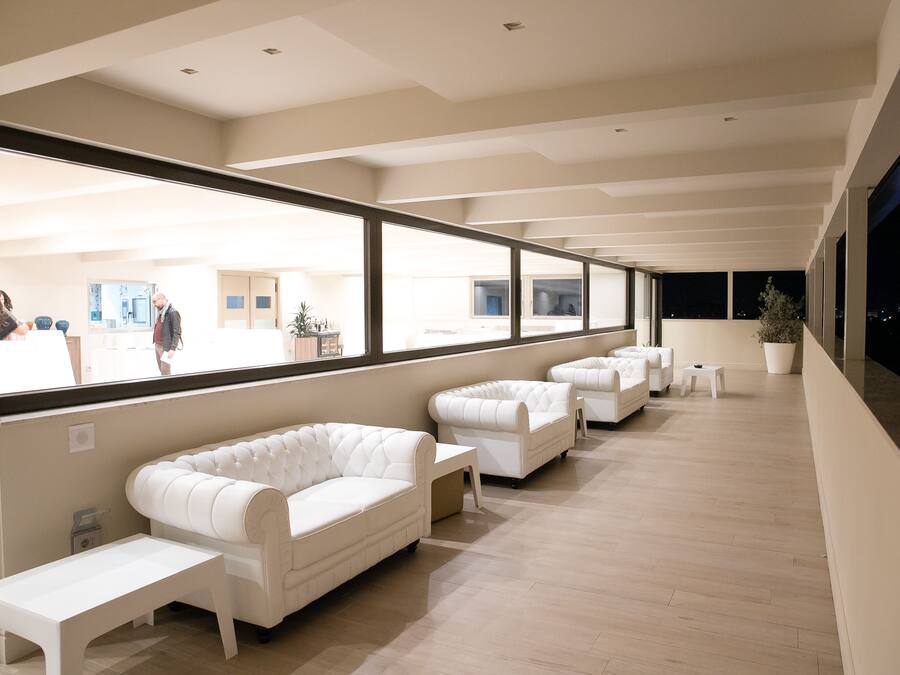 Un espace de détente moderne avec des canapés en cuir blanc et un plancher en bois, de grandes fenêtres offrant une vue sur la ville, et un balcon agrémenté de plantes en pot.