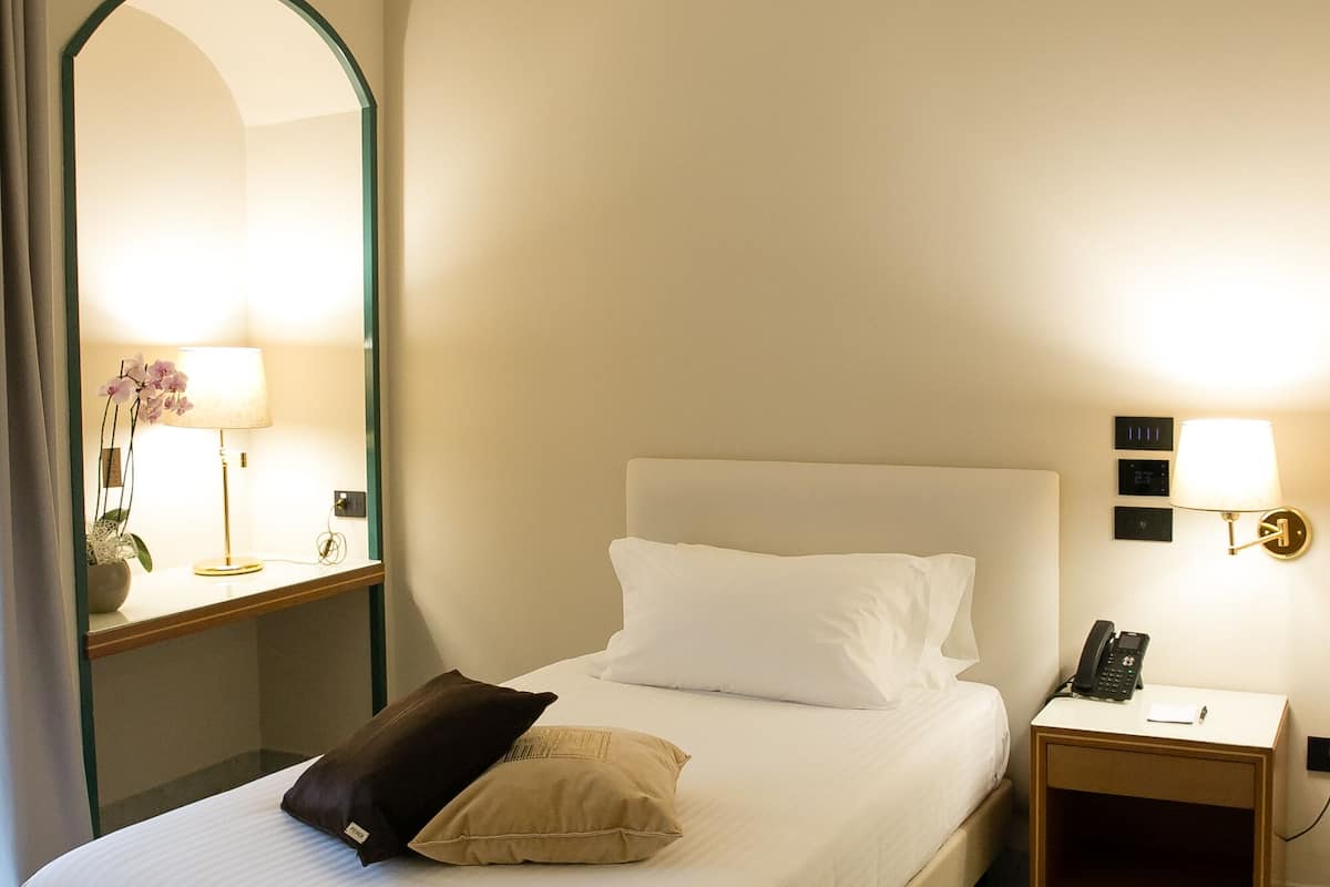 A neatly made bed with white linens and two pillows, a bedside table with a lamp, a phone, and a wall-mounted light fixture.