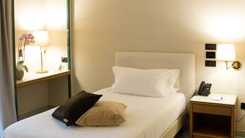 A neatly made bed with white linens and two pillows, a bedside table with a lamp, a phone, and a wall-mounted light fixture.
