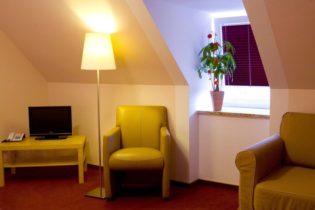Quarto pequeno e acolhedor com uma poltrona amarela, um sofá, uma TV em uma mesinha e um vaso de planta perto de uma janela.