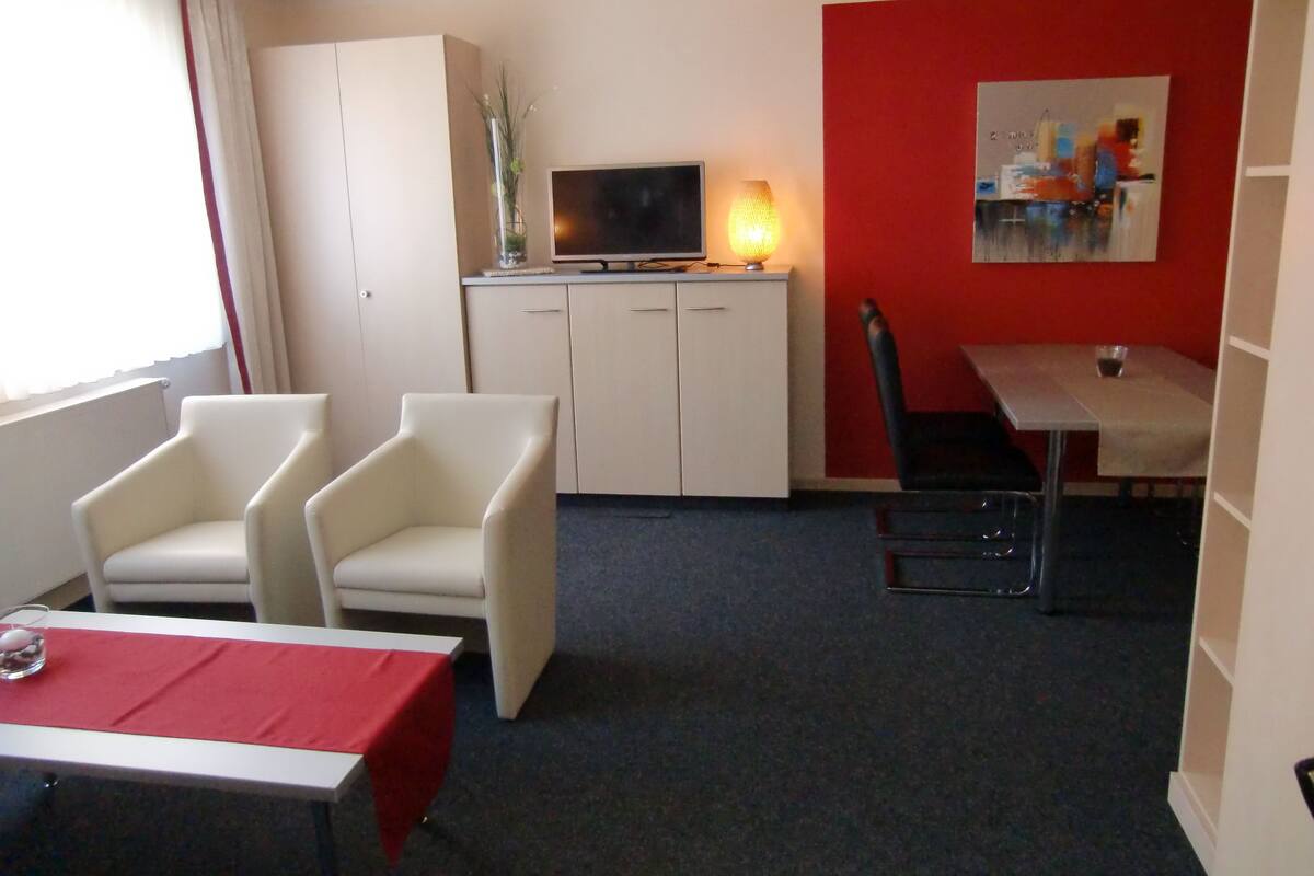 Quarto de hotel com televisão, pequena mesa de jantar, duas cadeiras, armário e parede vermelha.