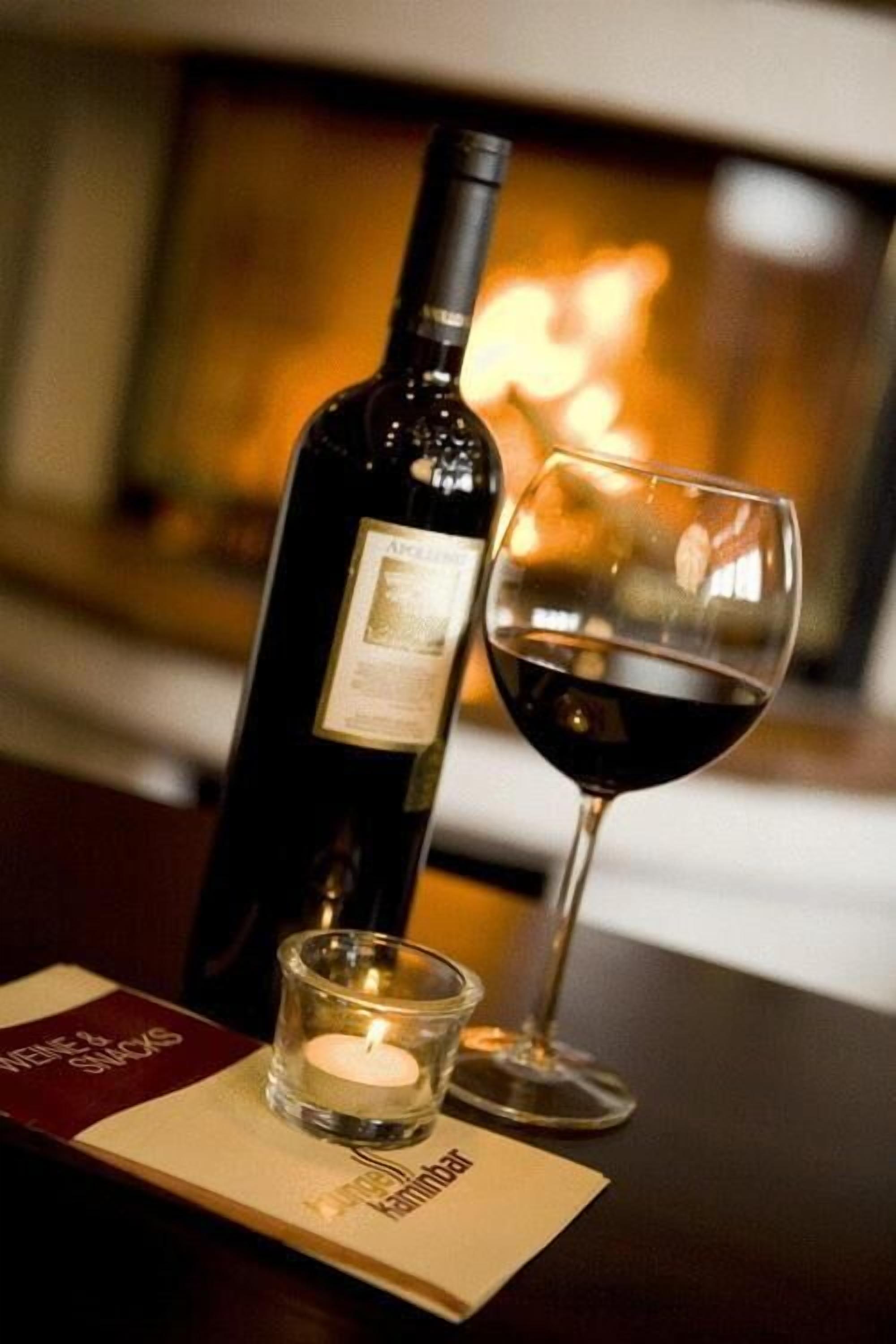 A bottle of wine and a glass on a table with a candle, a menu, and a fireplace in the background.