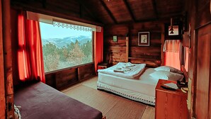 A wooden cabin bedroom with a bed, a bench, a nightstand, and a window with a view of the mountains. - Godence Hotel Restoran (Seferihisar)