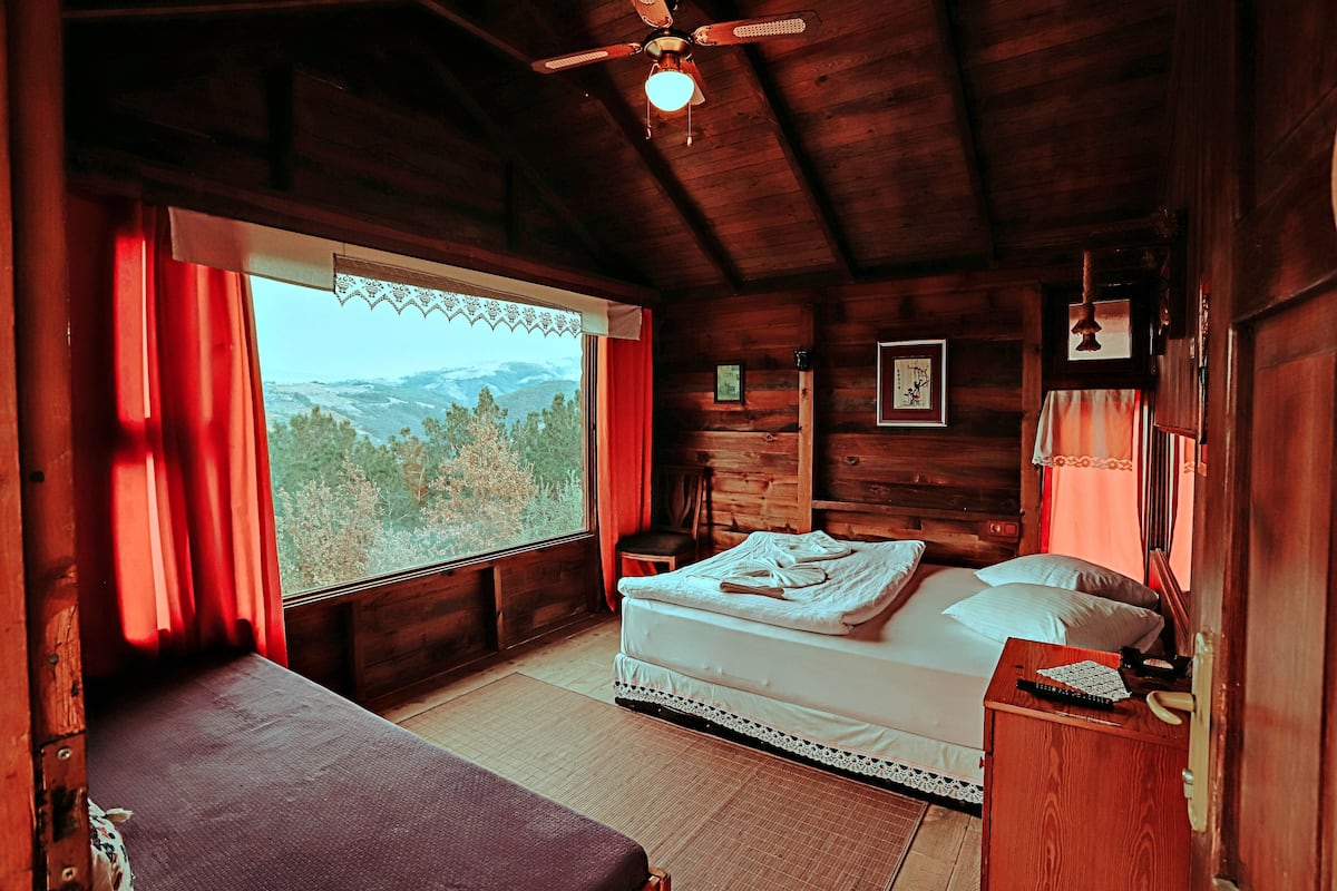 A wooden cabin bedroom with a bed, a bench, a nightstand, and a window with a view of the mountains.