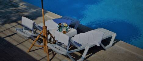 Un área junto a la piscina con sillones de descanso, una mesa y un paraguas rosa.