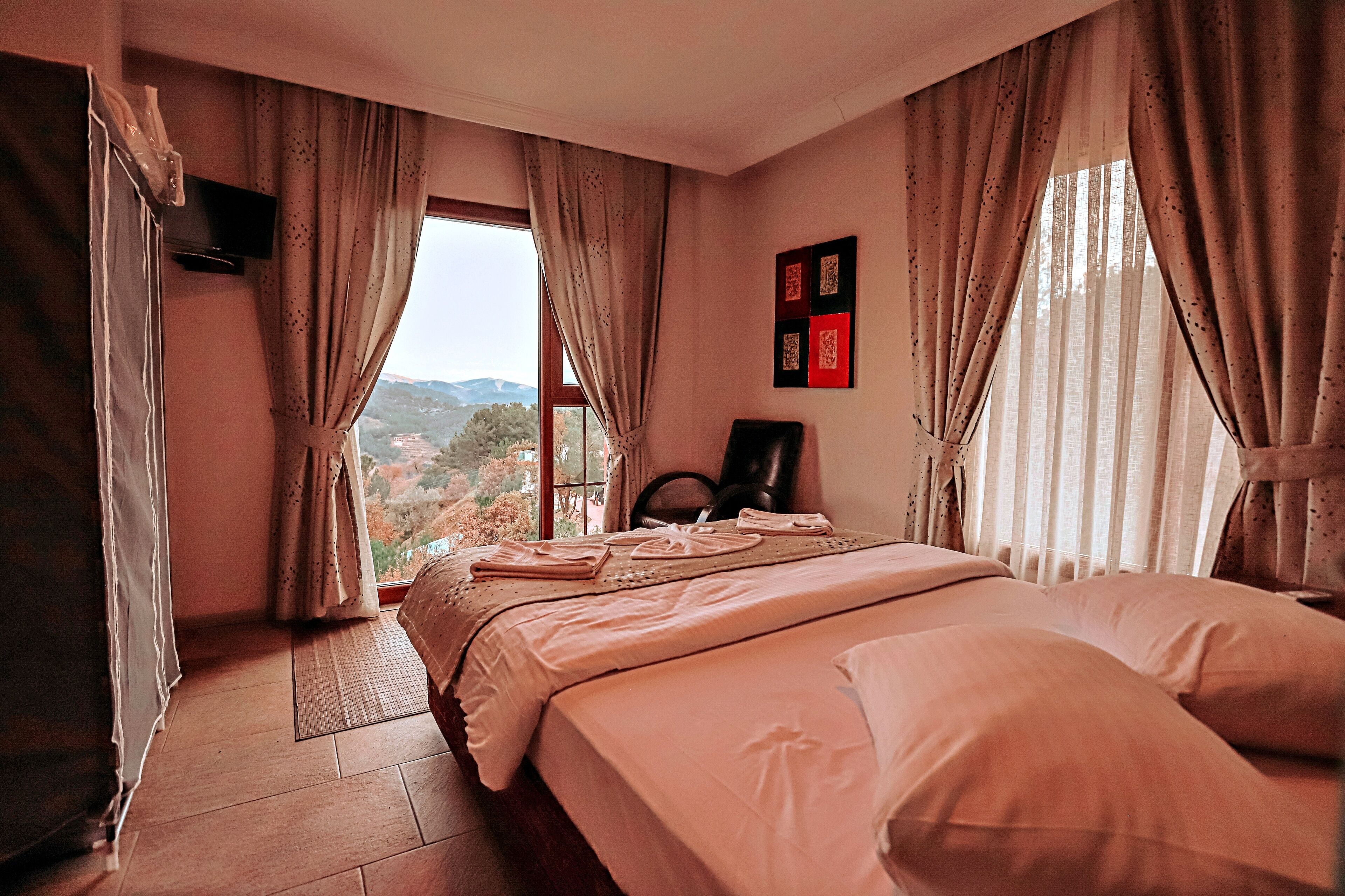 Un dormitorio con una cama grande, una silla, un televisor montado en la pared y un ventanal con cortinas.