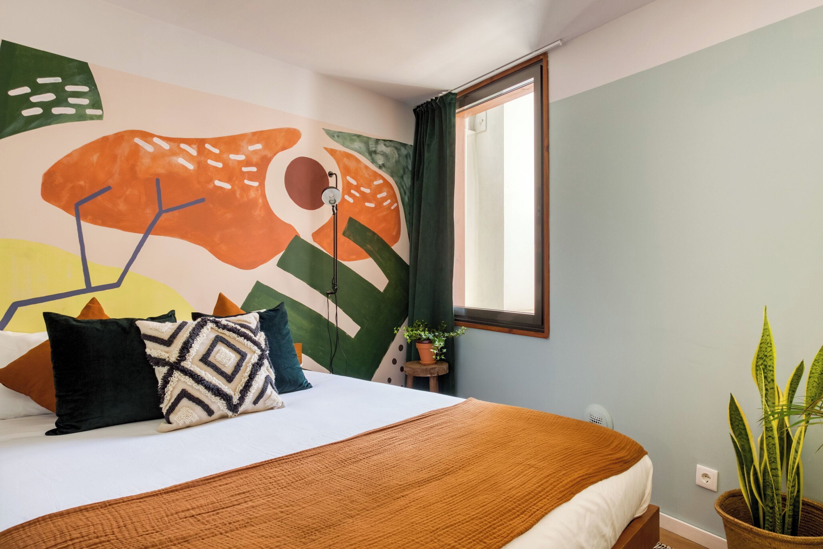 Un dormitori amb un llit, almoines, un mural a la paret, una finestra i una planta en test.