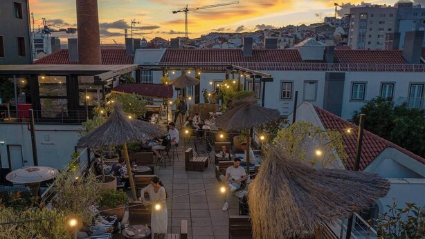 Een dakterras met terras, rieten parasols en lichtjes, met een stadsgezicht in de zonsondergang op de achtergrond.