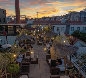 Un bar situé sur un toit, avec des places assises en plein air, des parasols en chaume et des guirlandes lumineuses, le tout surplombant un paysage urbain illuminé par le coucher du soleil.