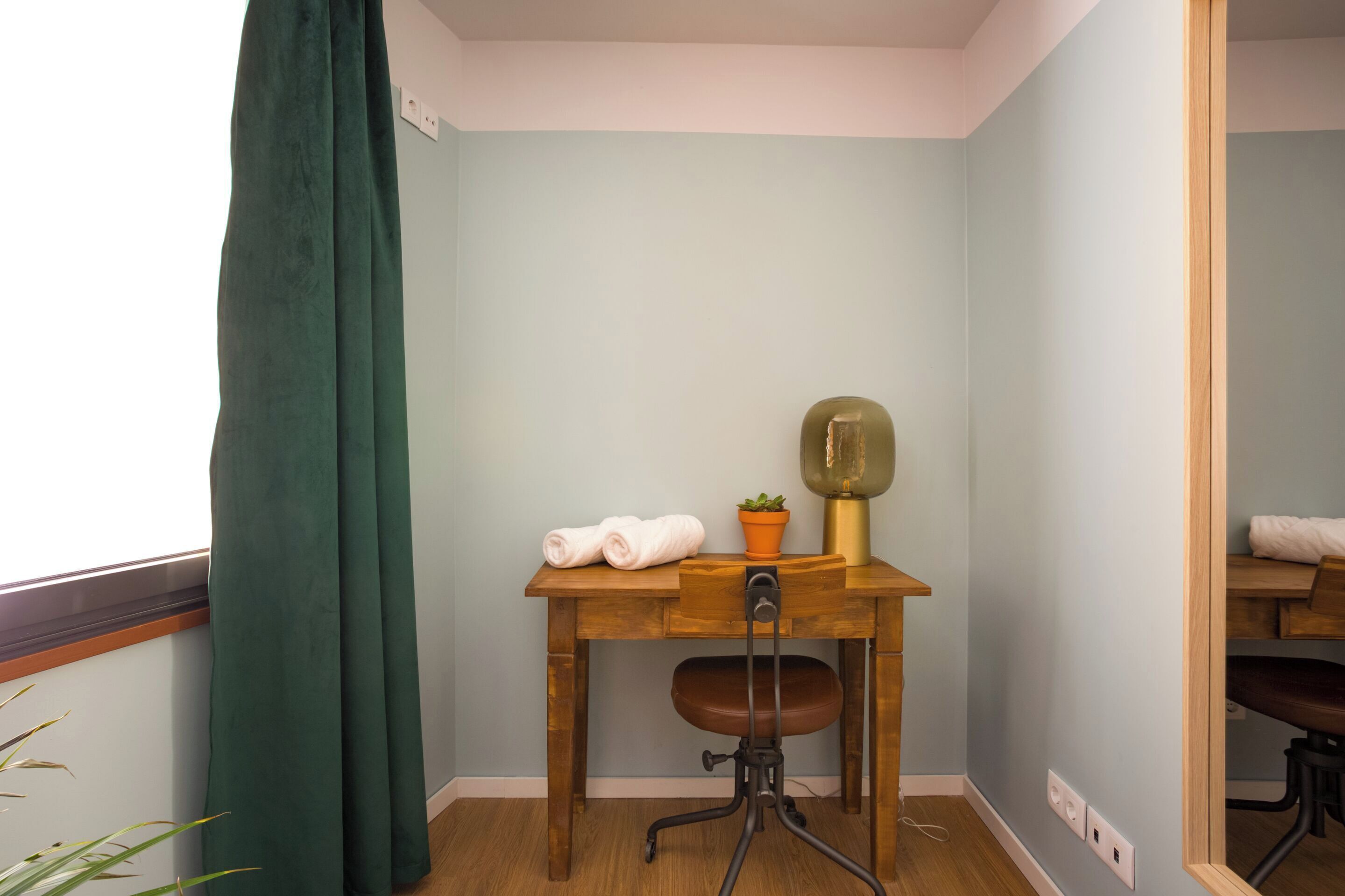 Una camera d'angolo con una scrivania in legno, una sedia, una pianta in vaso e una finestra con tenda verde.