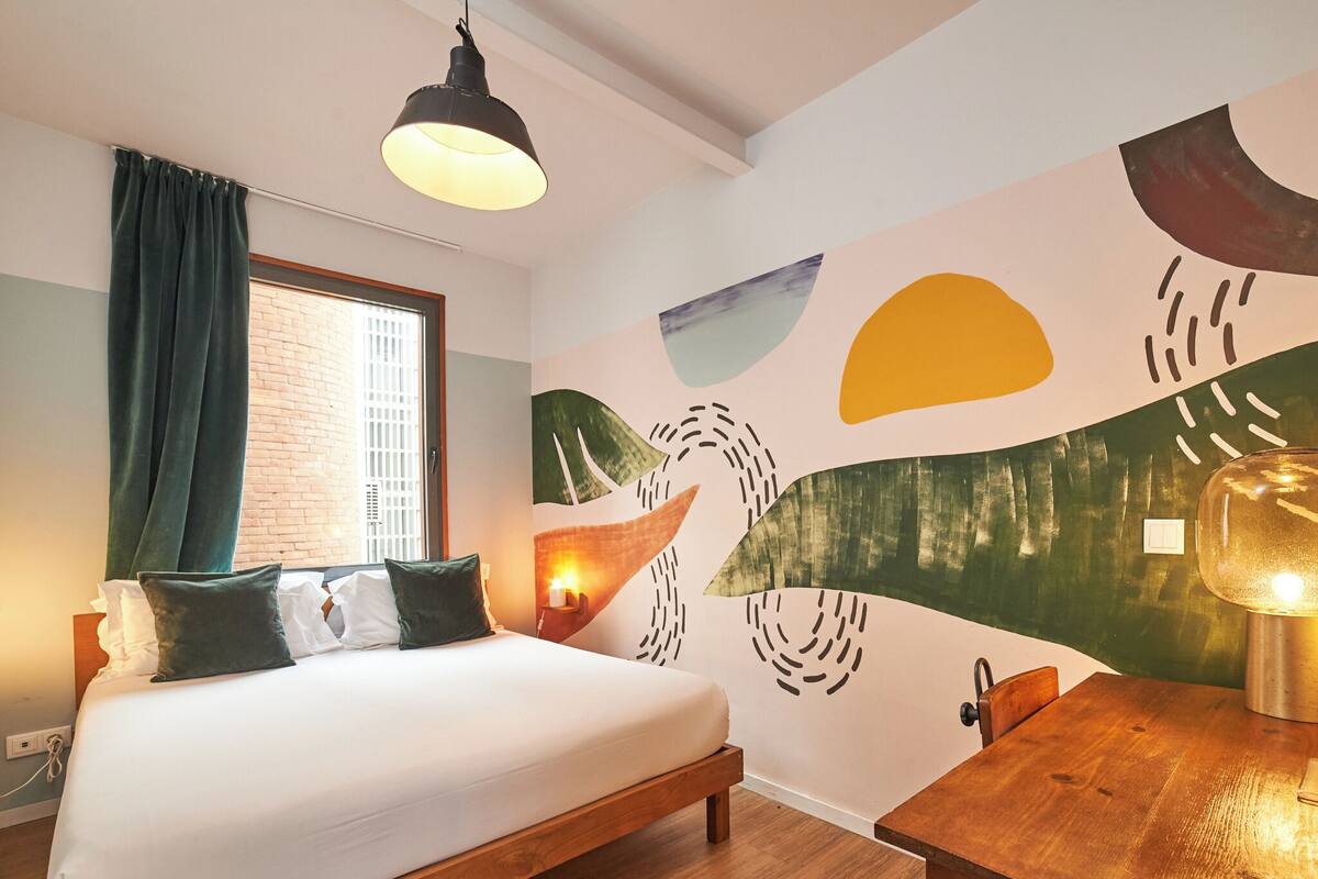 Ein Schlafzimmer mit einem Bett, einem Schreibtisch, einer Lampe, einem Fenster mit Vorhängen und einer Wand mit abstrakter Kunst.