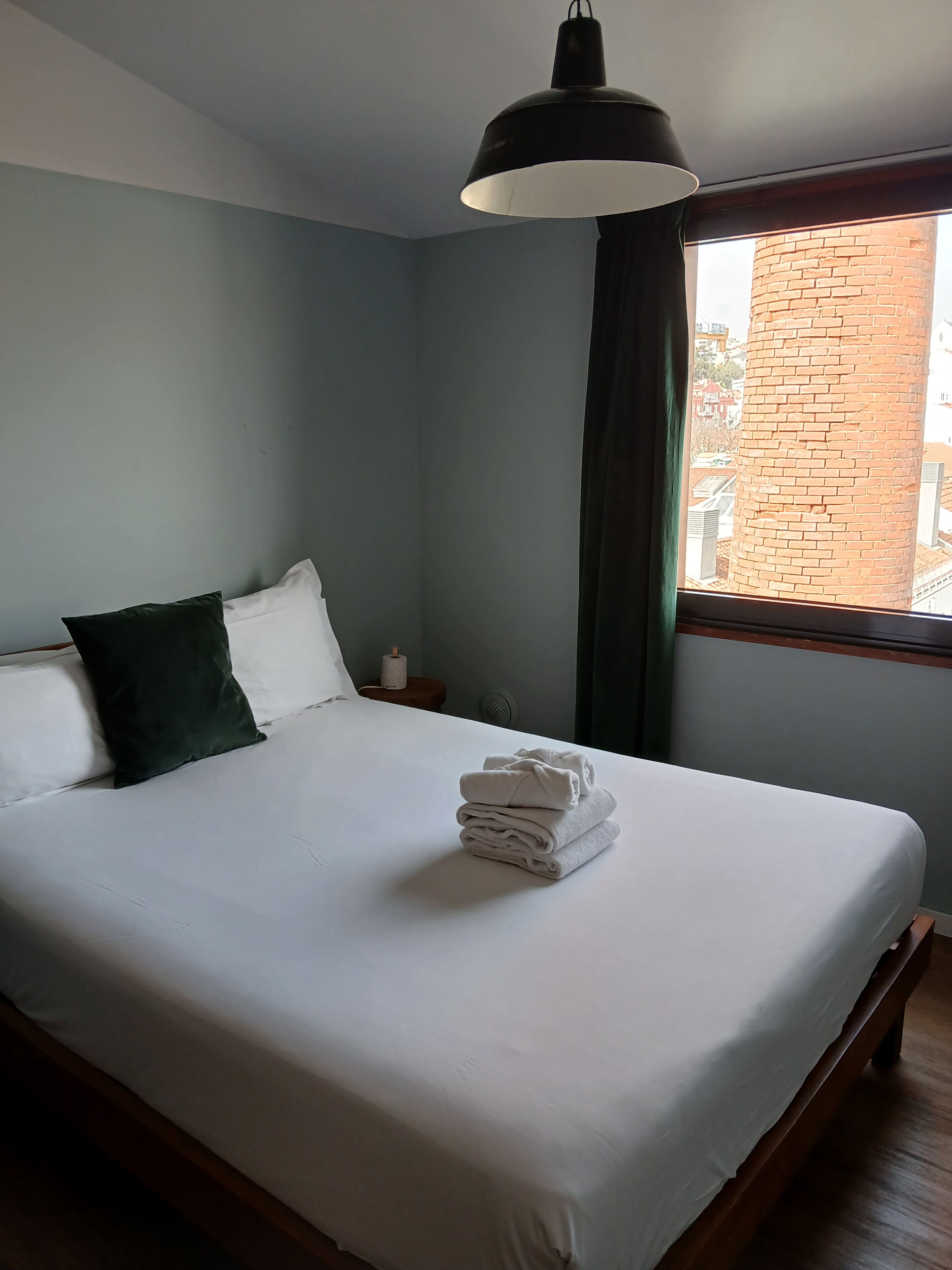 Un lit bien fait, avec du linge de lit blanc et une pile de serviettes pliées, un oreiller vert foncé, une lampe noire au plafond et une fenêtre donnant sur un mur de briques.