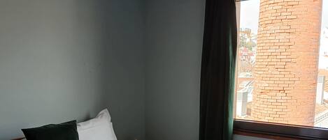 Un lit bien fait, avec du linge de lit blanc et une pile de serviettes pliées, un oreiller vert foncé, une lampe noire au plafond et une fenêtre donnant sur un mur de briques.
