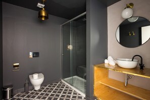 Een moderne badkamer met een douchecabine van glas, een wit toilet, een wastafel met een gouden aanrechtblad, en een ronde spiegel aan de muur.