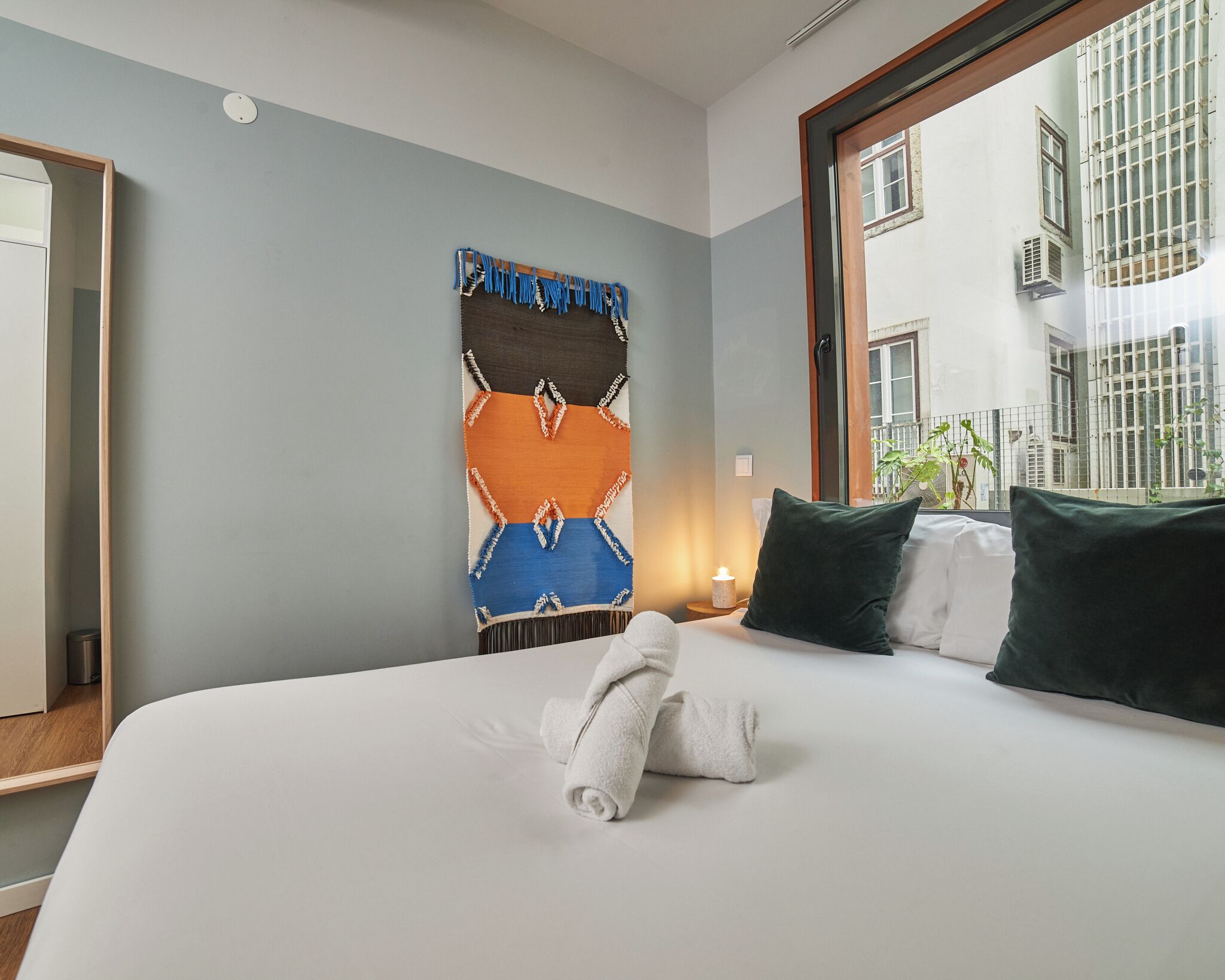 Habitación de hotel con cama, un tapiz en la pared, una ventana con vistas a edificios y una mesita de noche con una vela.