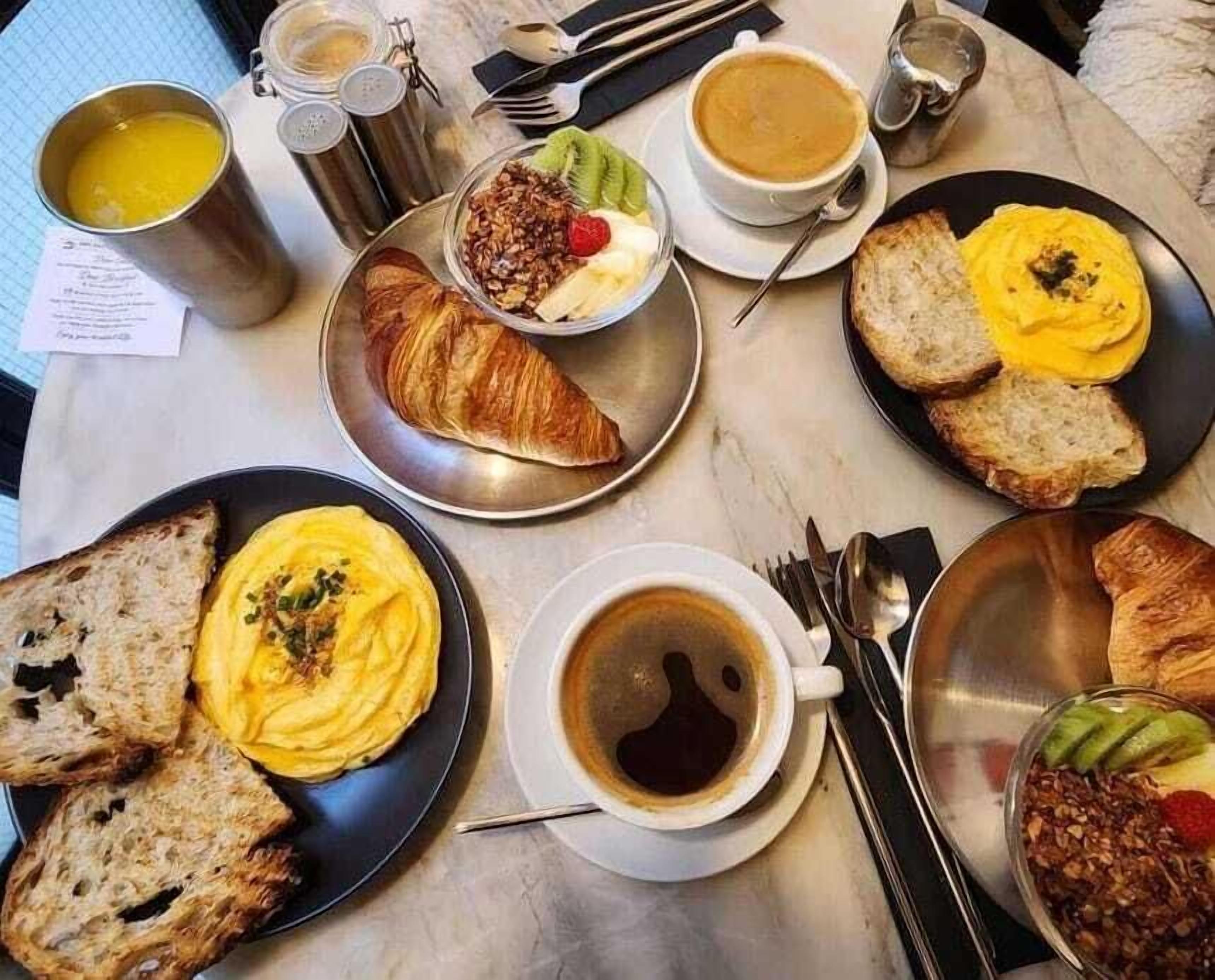 Morgenmadsbord med croissanter, ristet brød, æg, frugt og kaffe på et marmorbord.