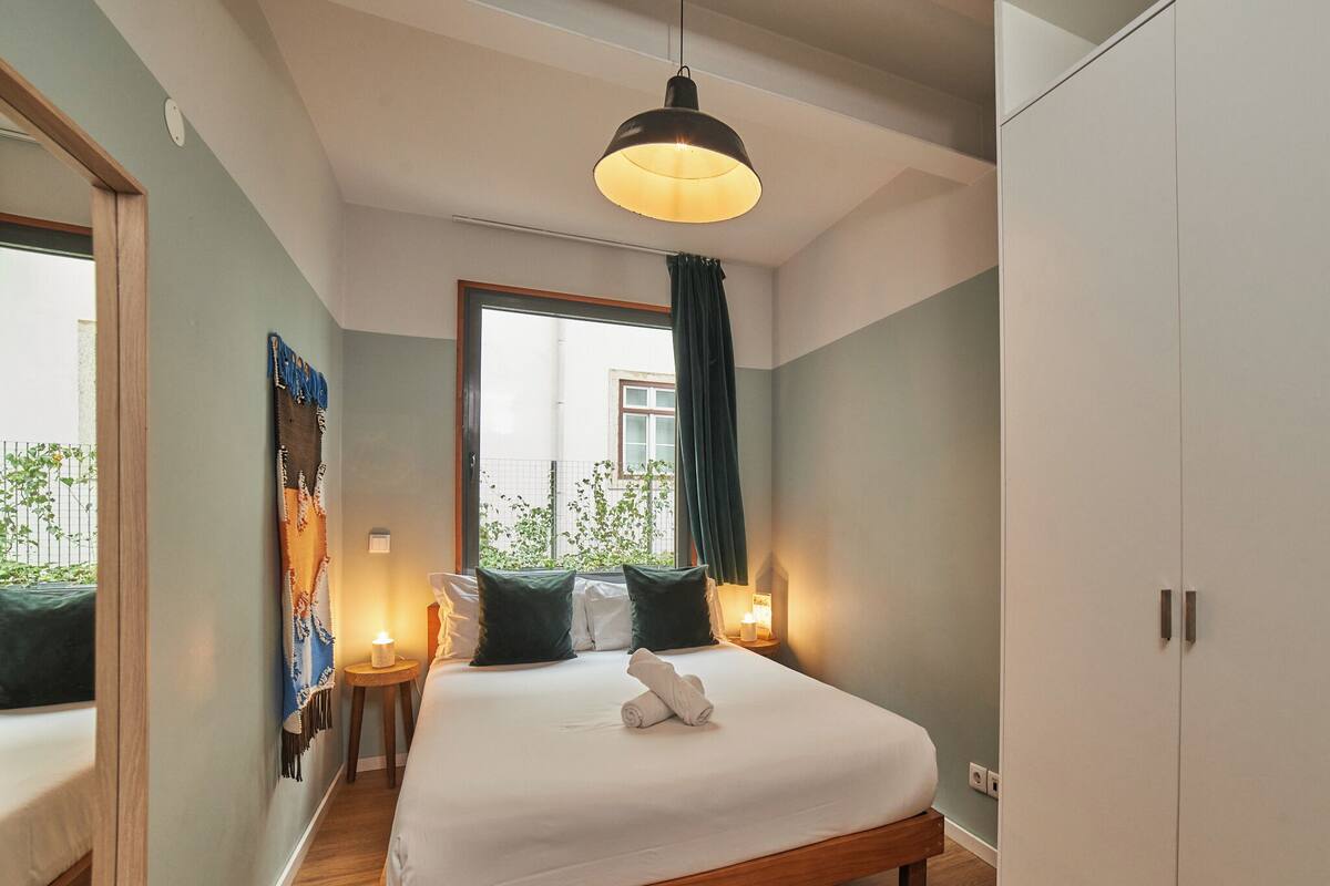 Ein Schlafzimmer mit einem großen Bett, Nachttischlampen, einem Fenster mit Vorhängen und Blick ins Grüne.