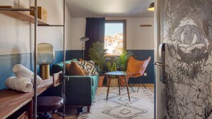 Een knus leefruimte met een bank, een fauteuil, een klein tafeltje, een lamp, een kamerplant en een uitzicht op gebouwen door een raam.