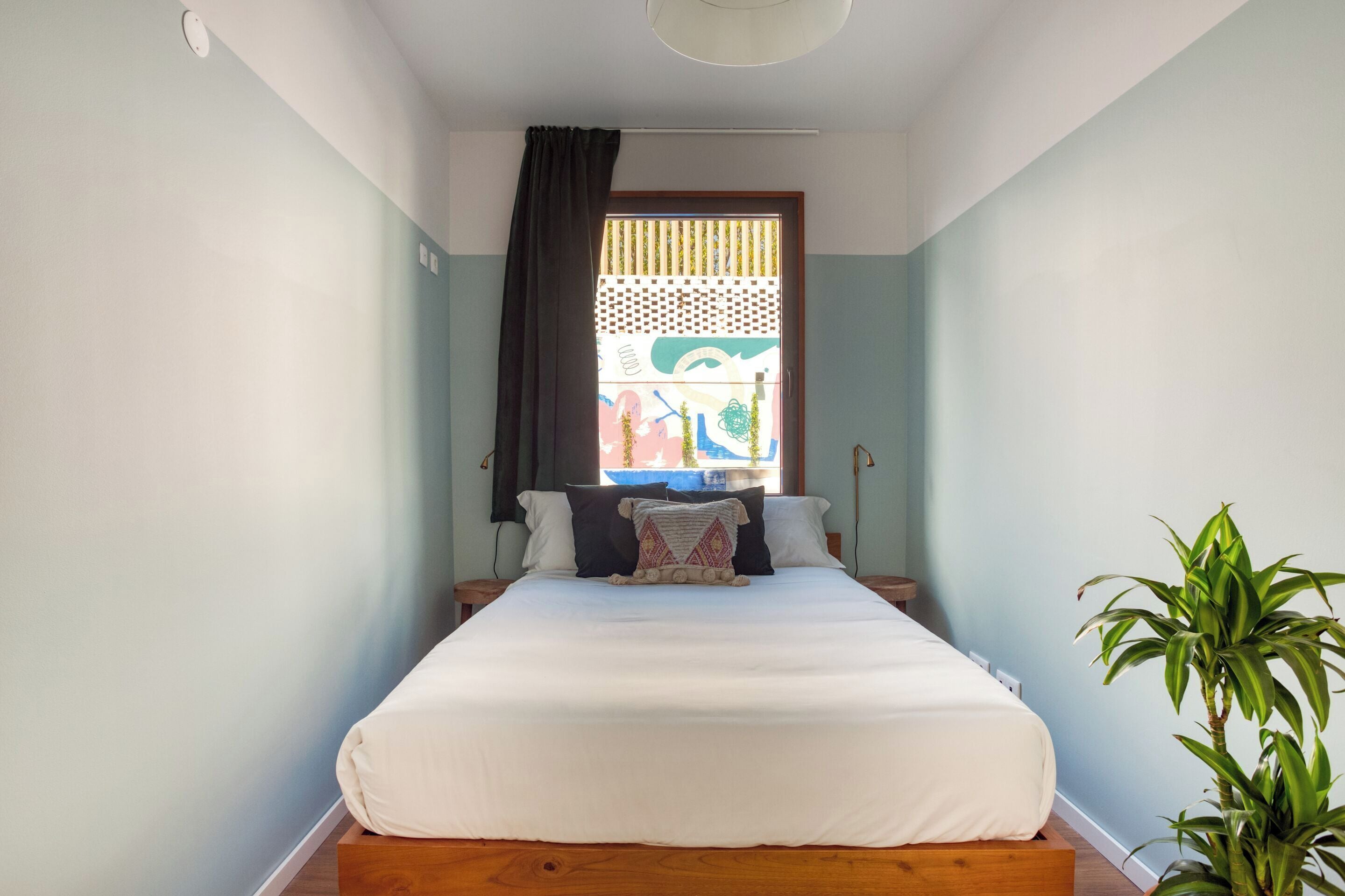 Un dormitorio con una cama, una ventana con vistas, una planta y una mesita de noche.