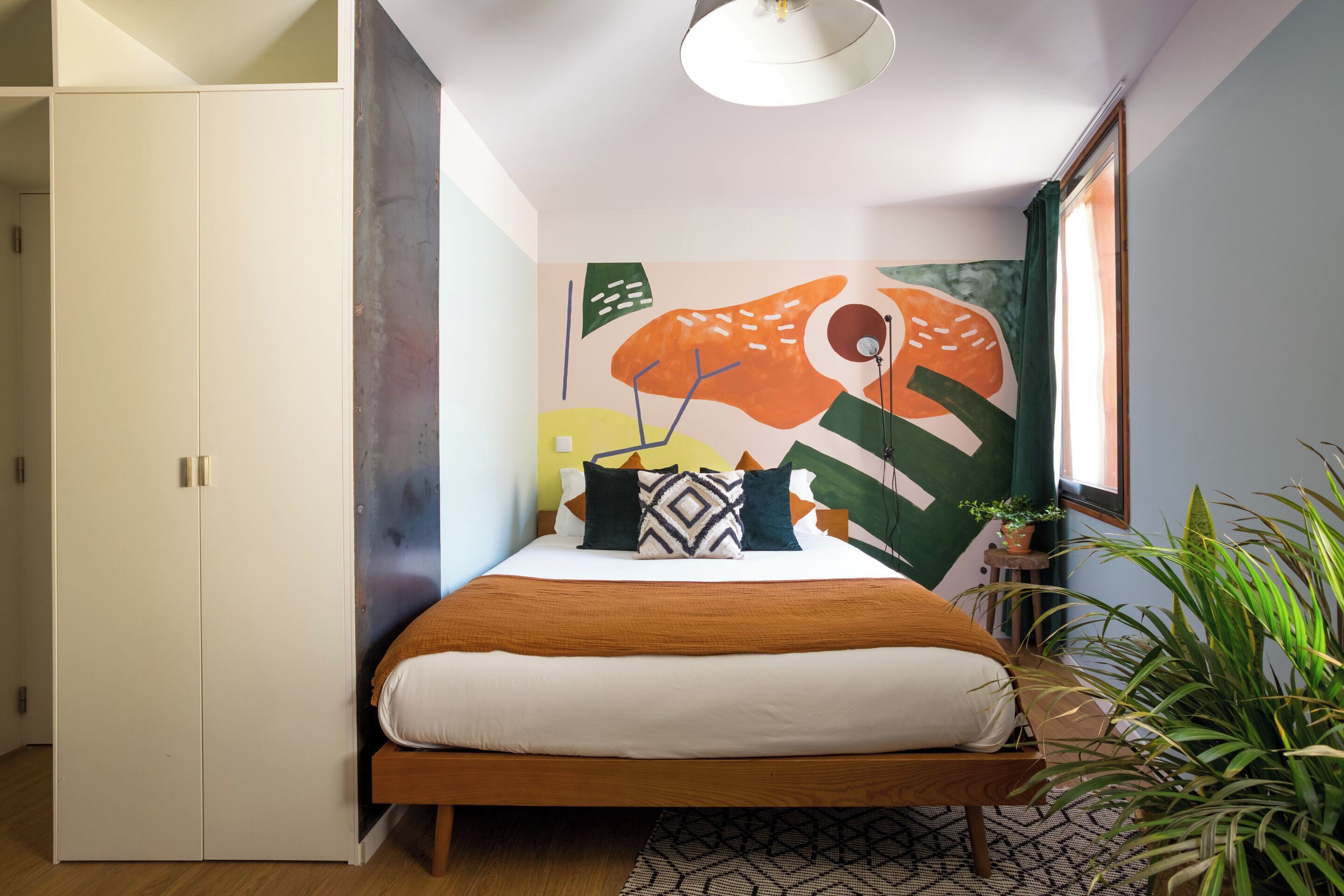 A bedroom with a bed, a wardrobe, a wall with abstract art, a window, and a plant.