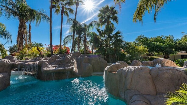 A pool area with clear blue water, rock formations, and palm trees under a bright sun.