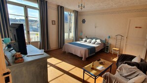 A room with a bed, a desk, a chair, a small table, and a window with curtains. - Joyeux Reveil (Courseulles-sur-Mer)