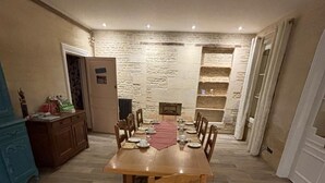 A dining area with a long wooden table set for six, surrounded by chairs. A stone wall with shelves is behind the table. - Joyeux Reveil (Courseulles-sur-Mer)