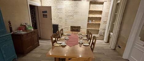 A dining area with a long wooden table set for six, surrounded by chairs. A stone wall with shelves is behind the table.
