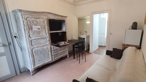 A room with a white sofa, a wooden cabinet, a small table with chairs, a TV, and a whiteboard. - Joyeux Reveil (Courseulles-sur-Mer)