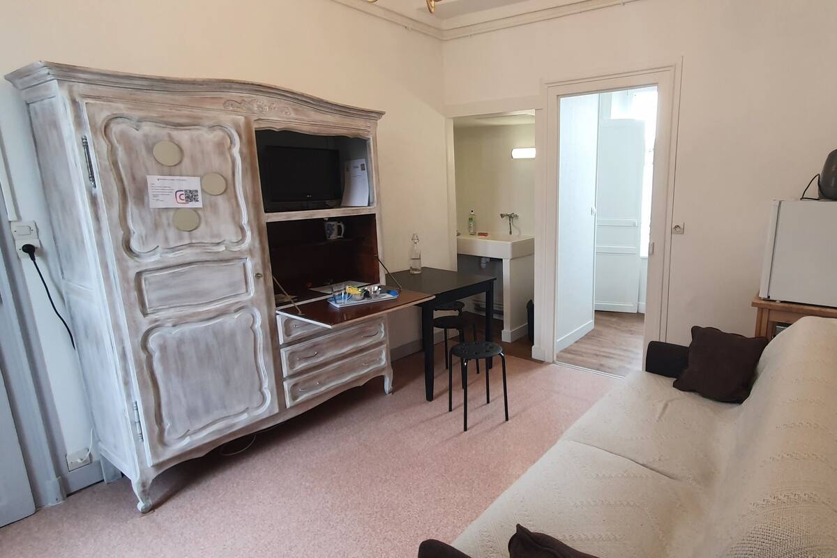 A room with a white sofa, a wooden cabinet, a small table with chairs, a TV, and a whiteboard.