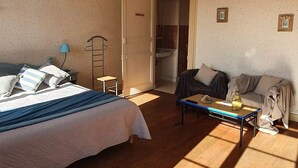 A bedroom with a bed, a small table, a sofa, and a bathroom visible through an open door. - Joyeux Reveil (Courseulles-sur-Mer)