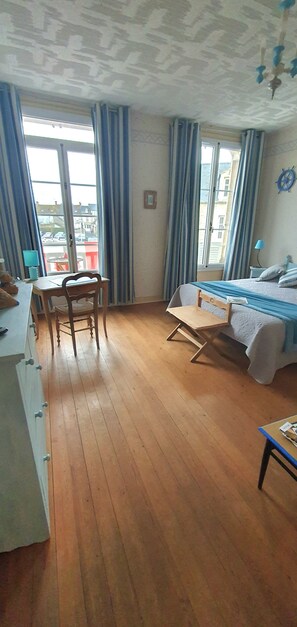 A room with a wooden floor, a bed with a blue blanket, a desk with a chair, and a window with curtains.