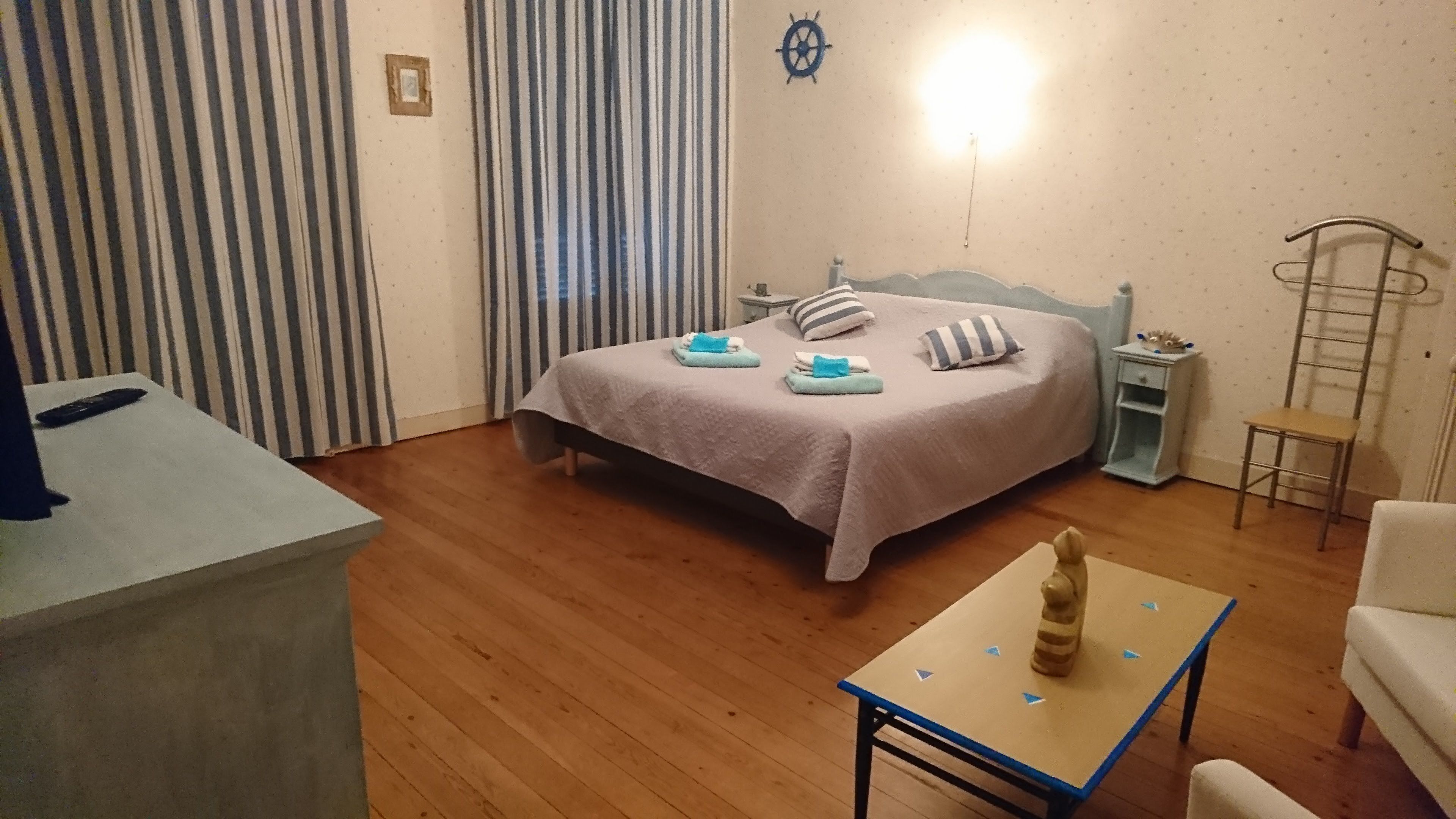 Un dormitorio con cama, mesita de noche, silla, una pequeña mesa y cortina.