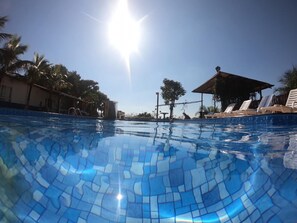 Una piscina azul cristalina con diseños de mosaicos, rodeada de palmeras y una zona sombreada con sillones de descanso.