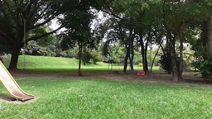 Un parc doté d’une aire de jeux, comprenant un toboggan et des balançoires, entouré d’arbres et de pelouse.