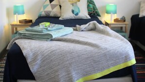 A neatly made bed with a grey blanket, a folded towel, and a small green item on the bed. There are two bedside tables with lamps, a patterned pillow, and a geometric pillow on the bed.