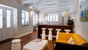 A modern kitchen with white cabinetry, a wooden island with stools, and a yellow sofa in the living area. - Villa Amalie (Willemstad)
