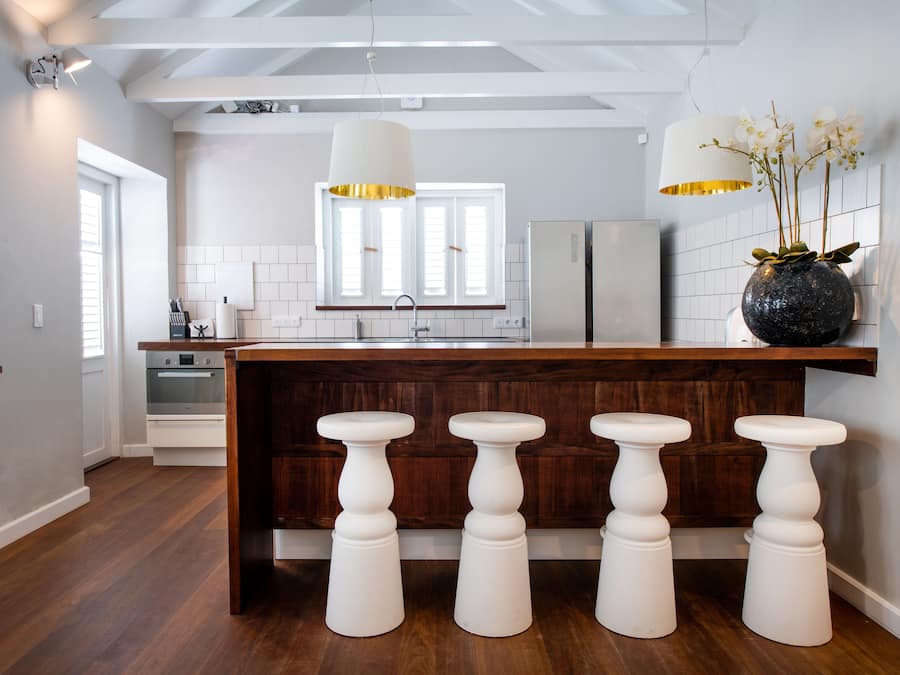 Una cocina moderna con una isla central, taburetes de madera, armarios blancos y un ventanal grande.