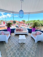 Un espacio de estar al aire libre techado, con sillas y sofá blancos, baldosas de suelo con diseño y vistas a la ciudad.