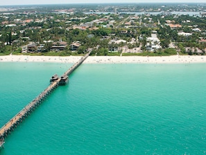 An aerial view of a coastal area with a long pier extending into turquoise waters, surrounded by residential buildings and a sandy beach. - Cozy apart in a quiet neighborhood, with beautiful views. (NAPLES)