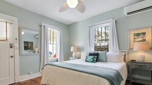 Quarto com uma cama, duas mesas de cabeceira, um ventilador de teto, uma janela com cortinas e um ar-condicionado fixado na parede.