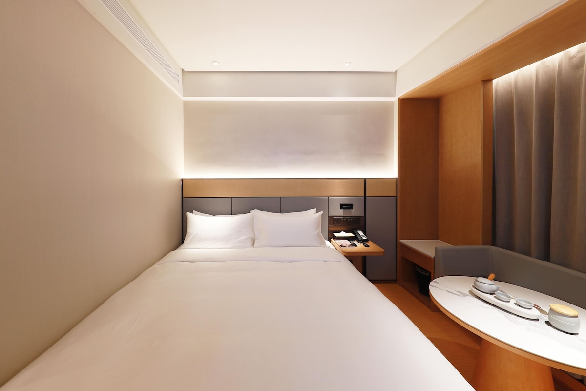 Una habitación de hotel moderna con una cama bien hecha, una mesita de noche con lámpara y una pequeña zona de estar con mesa.