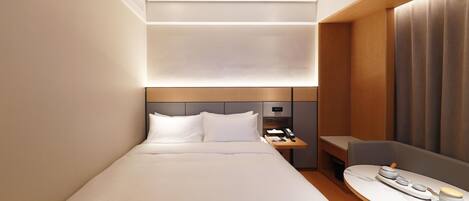 A modern hotel room with a neatly made bed, a bedside table with a lamp, and a small seating area with a table.
